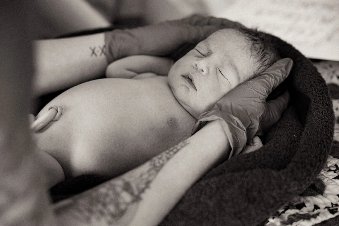 A newborn minutes old sleeps with eyes closed, cradled in the midwife's gloved hands, in black and white