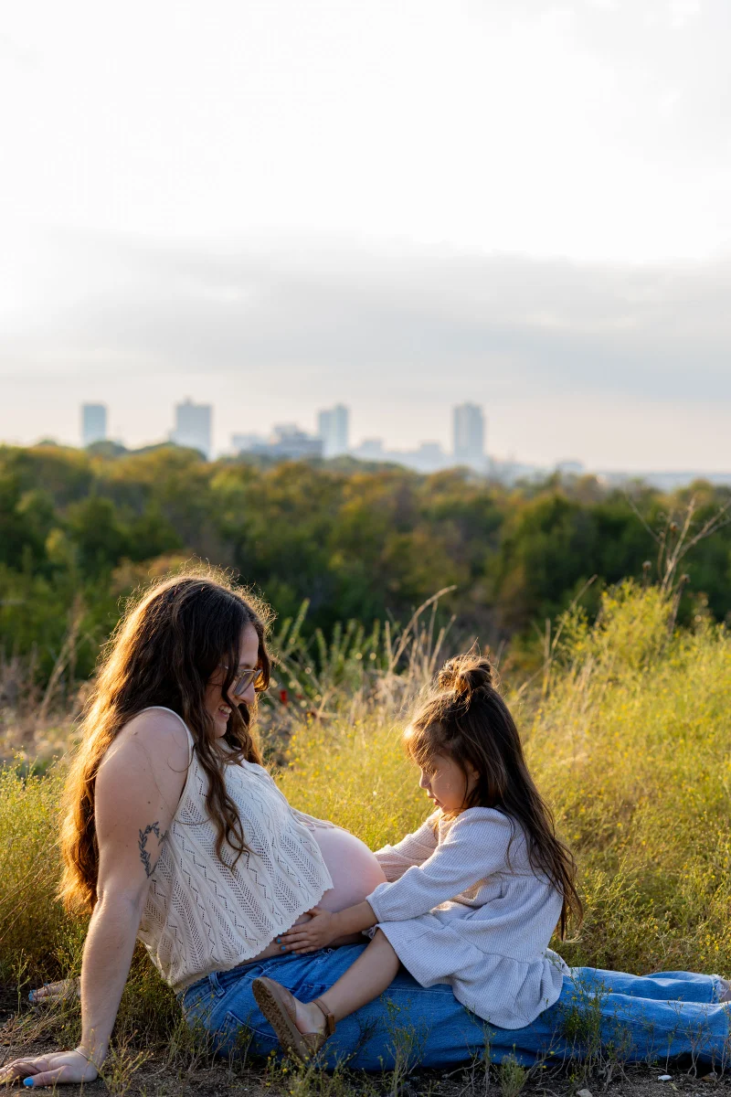 A pregnant mother sits in tall grass while her daughter rests both hands on her belly, a city skyline behind them
