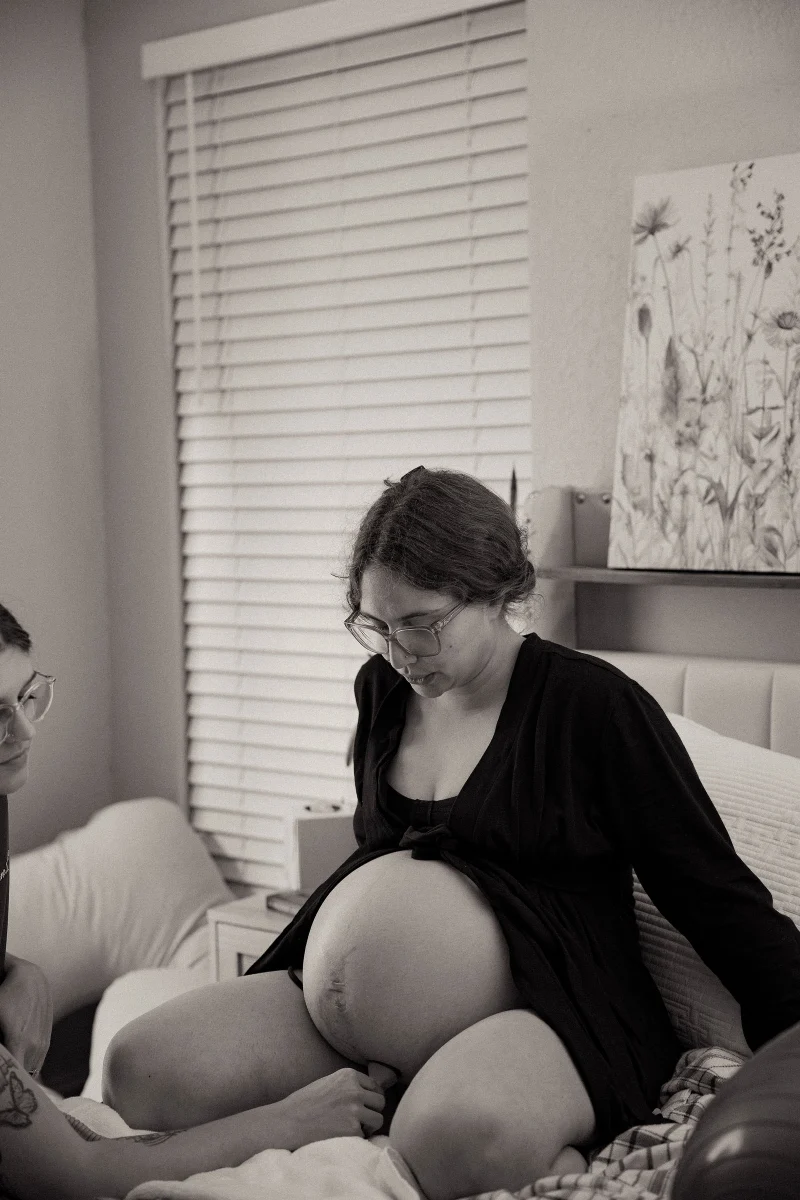 A laboring mother in glasses sits on her bed at home and looks down at her belly, in black and white