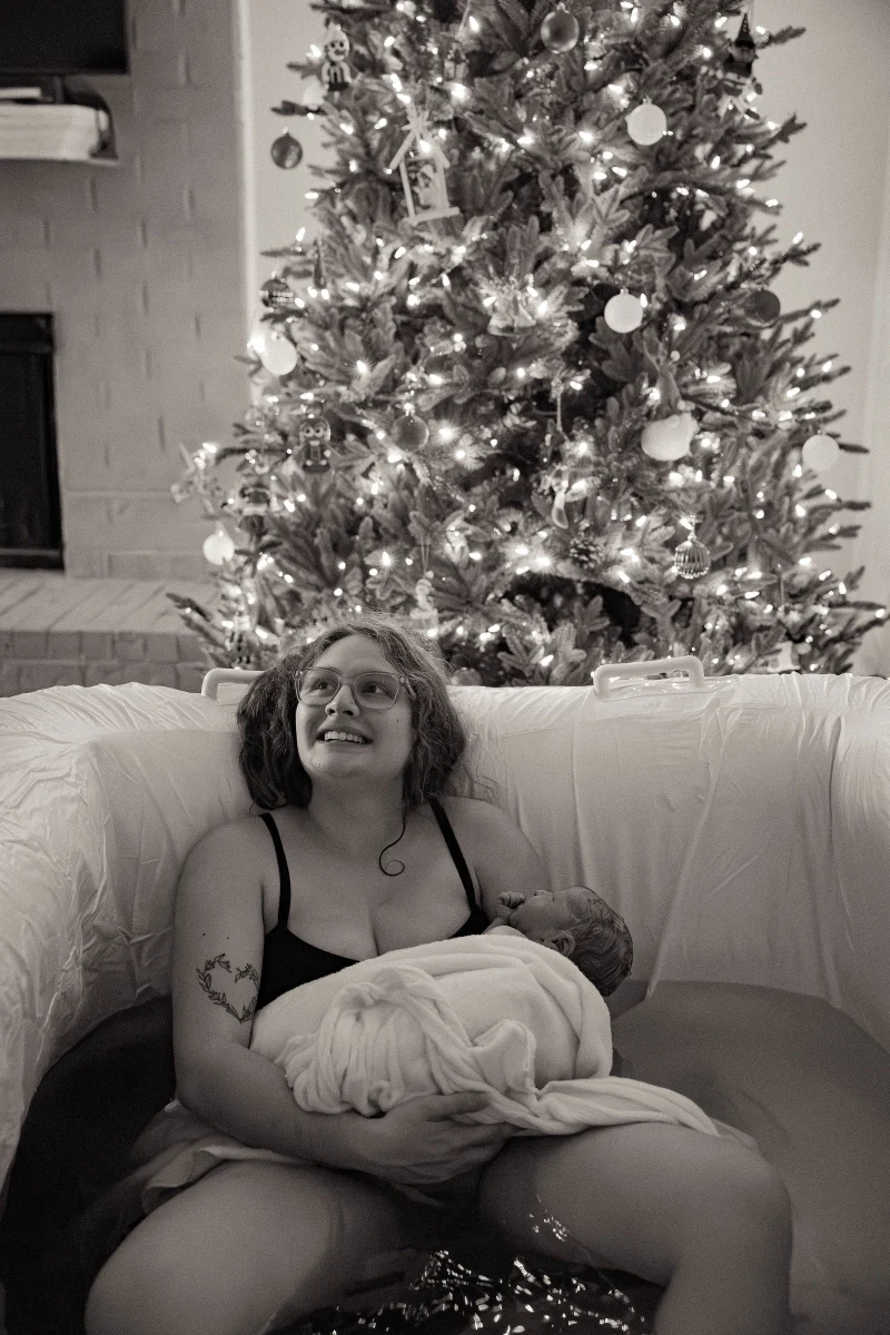 A mother smiling in a birth pool with her newborn wrapped in a towel, a lit Christmas tree behind her, in black and white